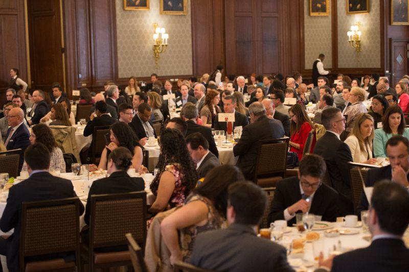 Large group of professionally dressed individuals seated at round tables during a formal banquet or conference in an ornate dining hall with wood-paneled walls and chandeliers.
